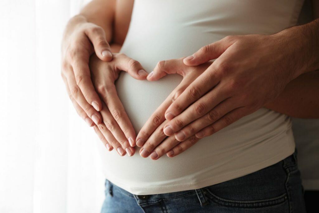 Dłonie kobiety w ciąży i mężczyzny trzymają za brzuch, palce układają się w kształt serca. Płodność kobiety.